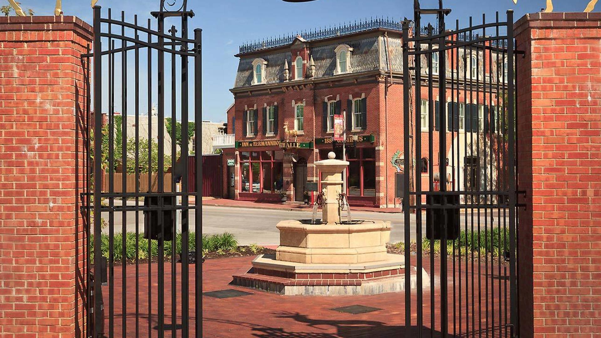 A wrought-iron gate frames a brick plaza with a fountain and a historic building in the background.