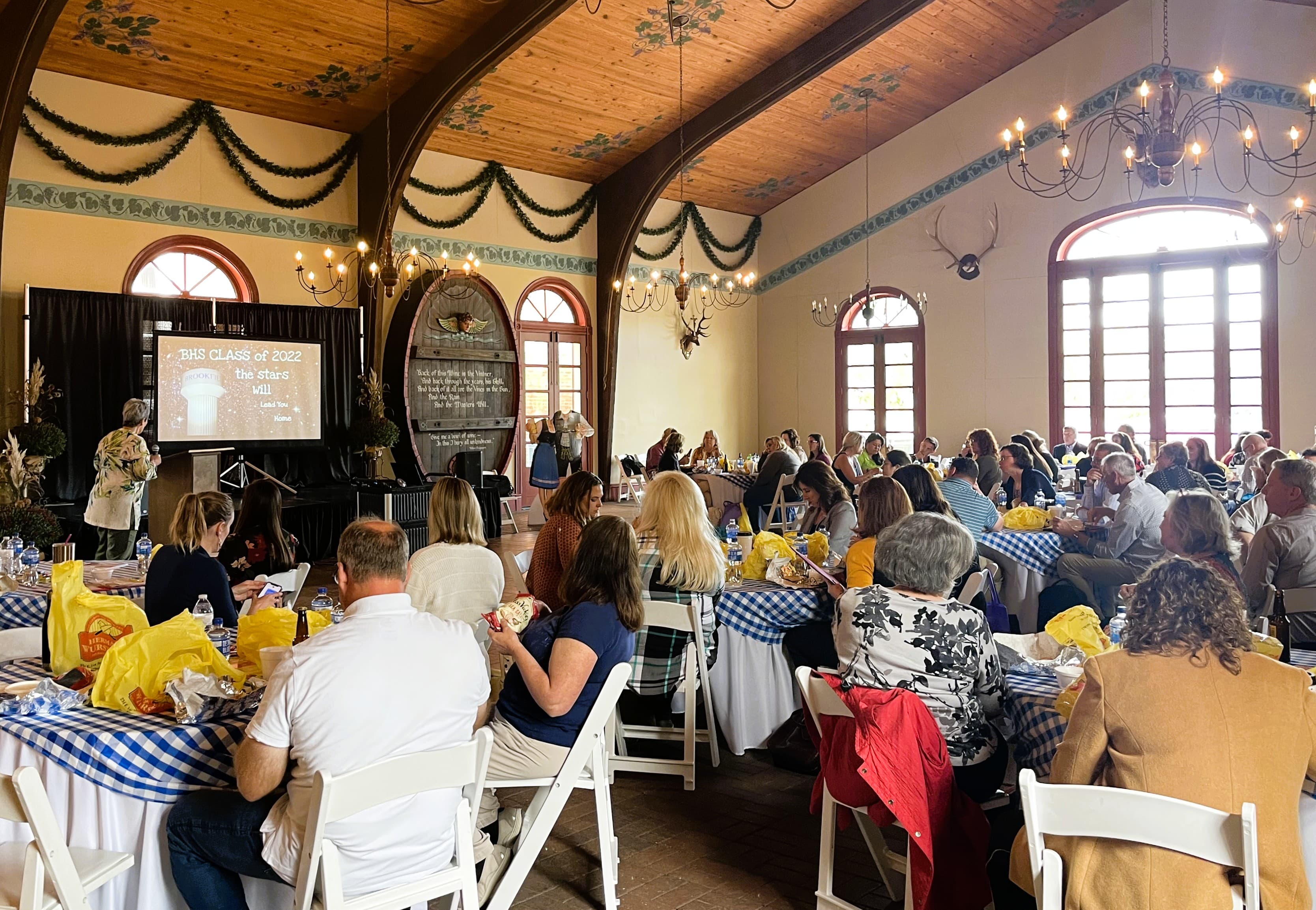 A large group of people gathers around tables in a decorated hall for an event featuring a presentation on a screen.