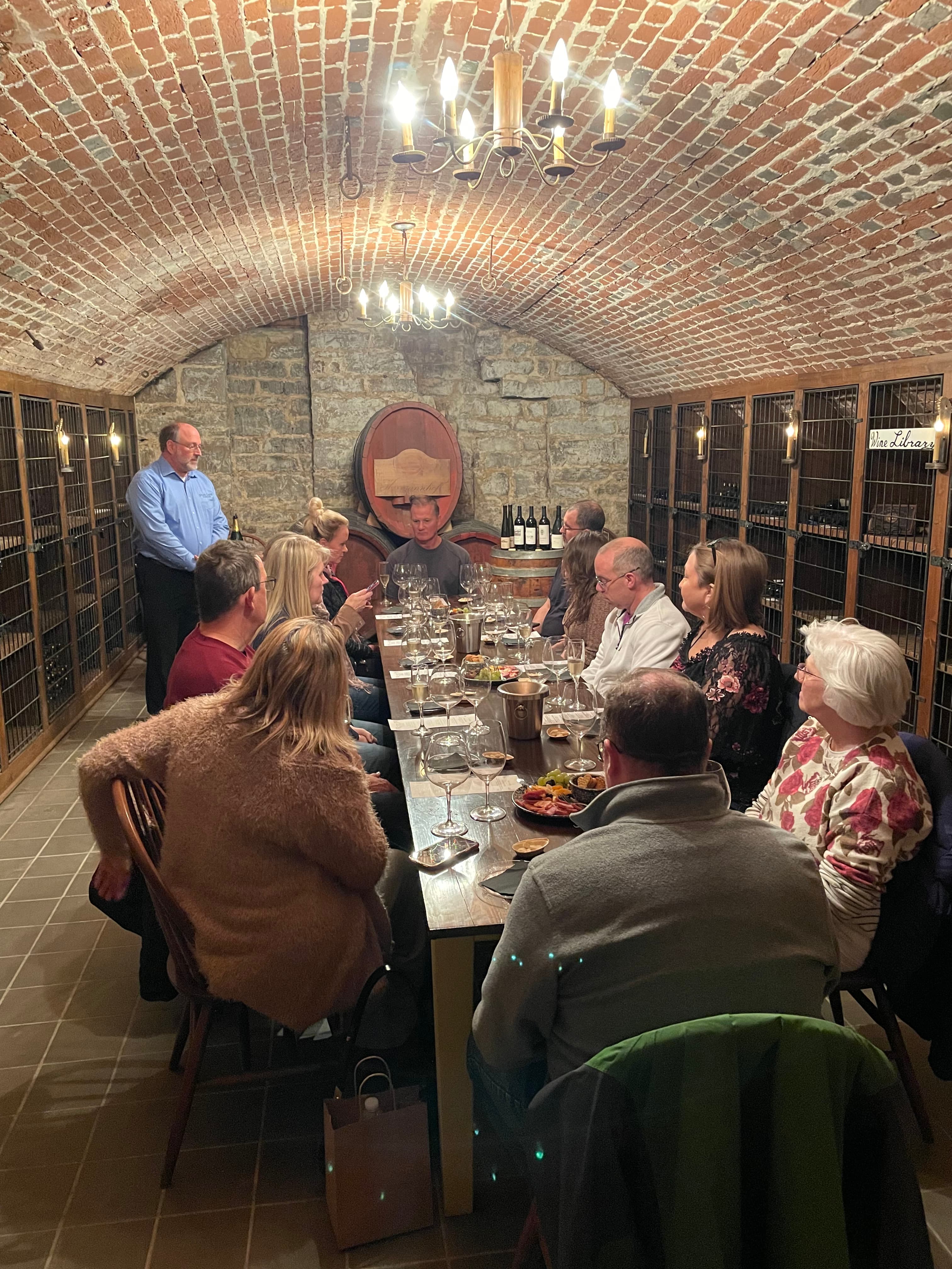 A group of diners sits around a long table in a rustic wine cellar, attentively listening to a speaker.