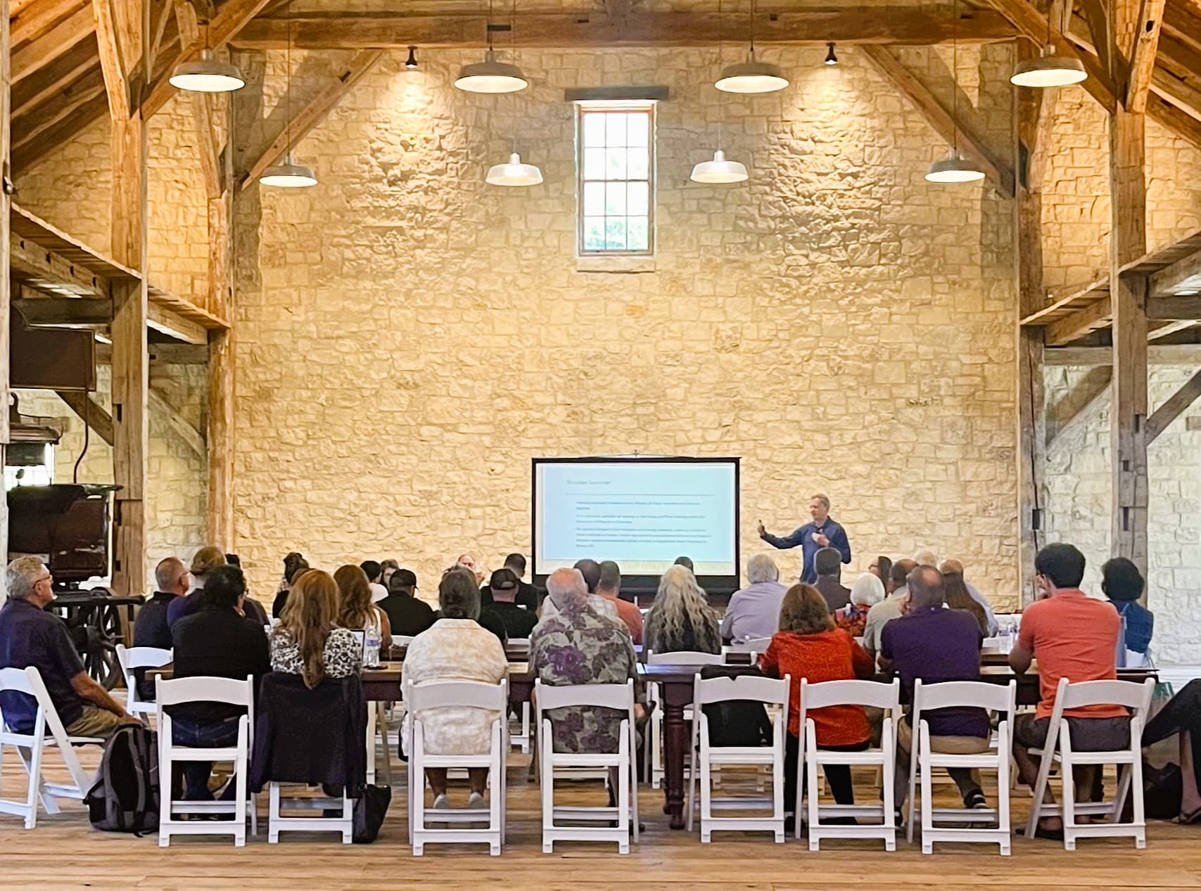 A speaker presents to an audience seated at long tables in a rustic hall.