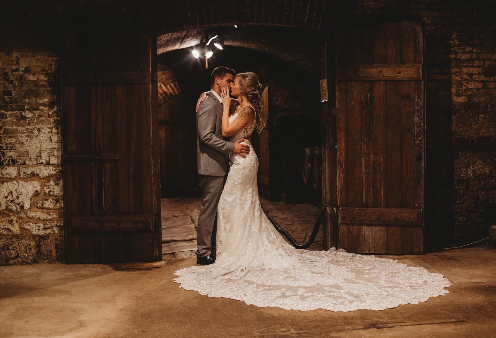 A couple shares a romantic kiss in a rustic venue, surrounded by wooden doors and brick walls.