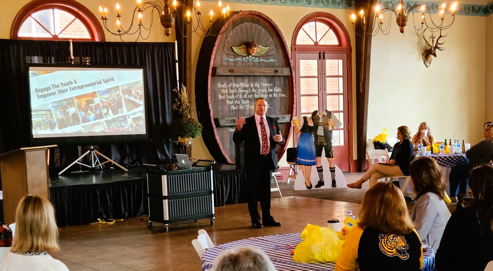 A speaker presents to an audience in a decorated hall with a large screen displaying the text "Engage The Youth & Empower Their Entrepreneurial Spirit."