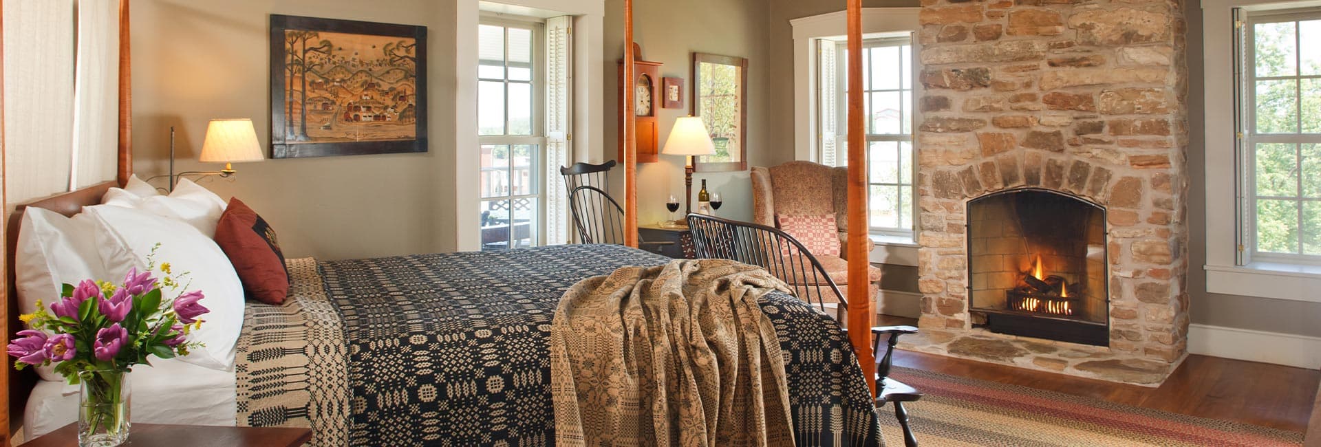 A cozy bedroom featuring a large bed, stone fireplace, and natural light from surrounding windows.