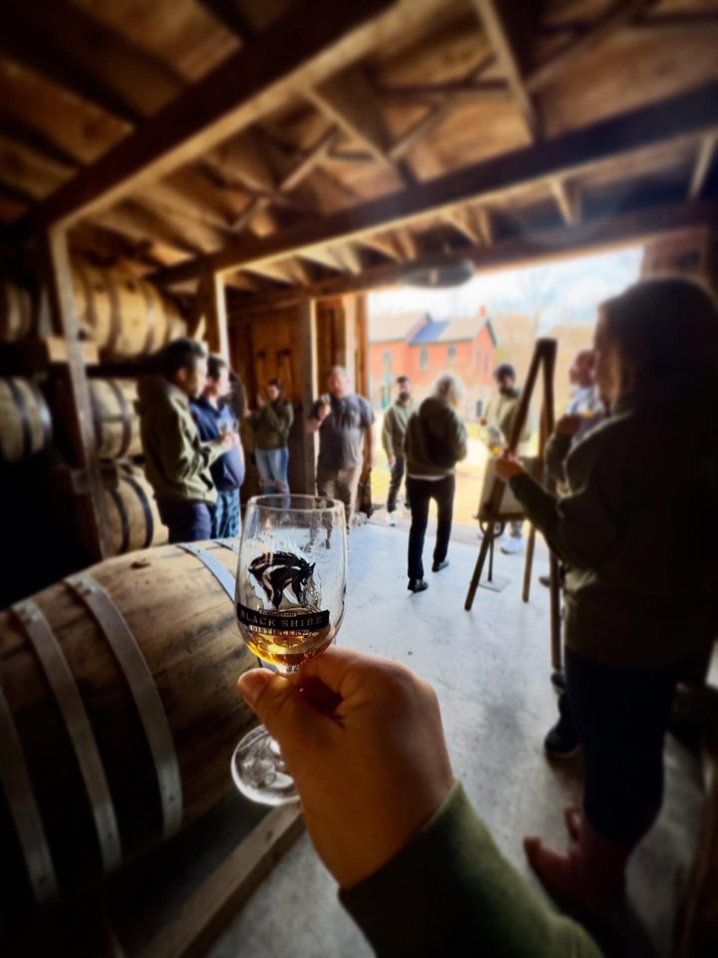 A hand holds a glass of whiskey in a rustic barn filled with people engaged in conversation.