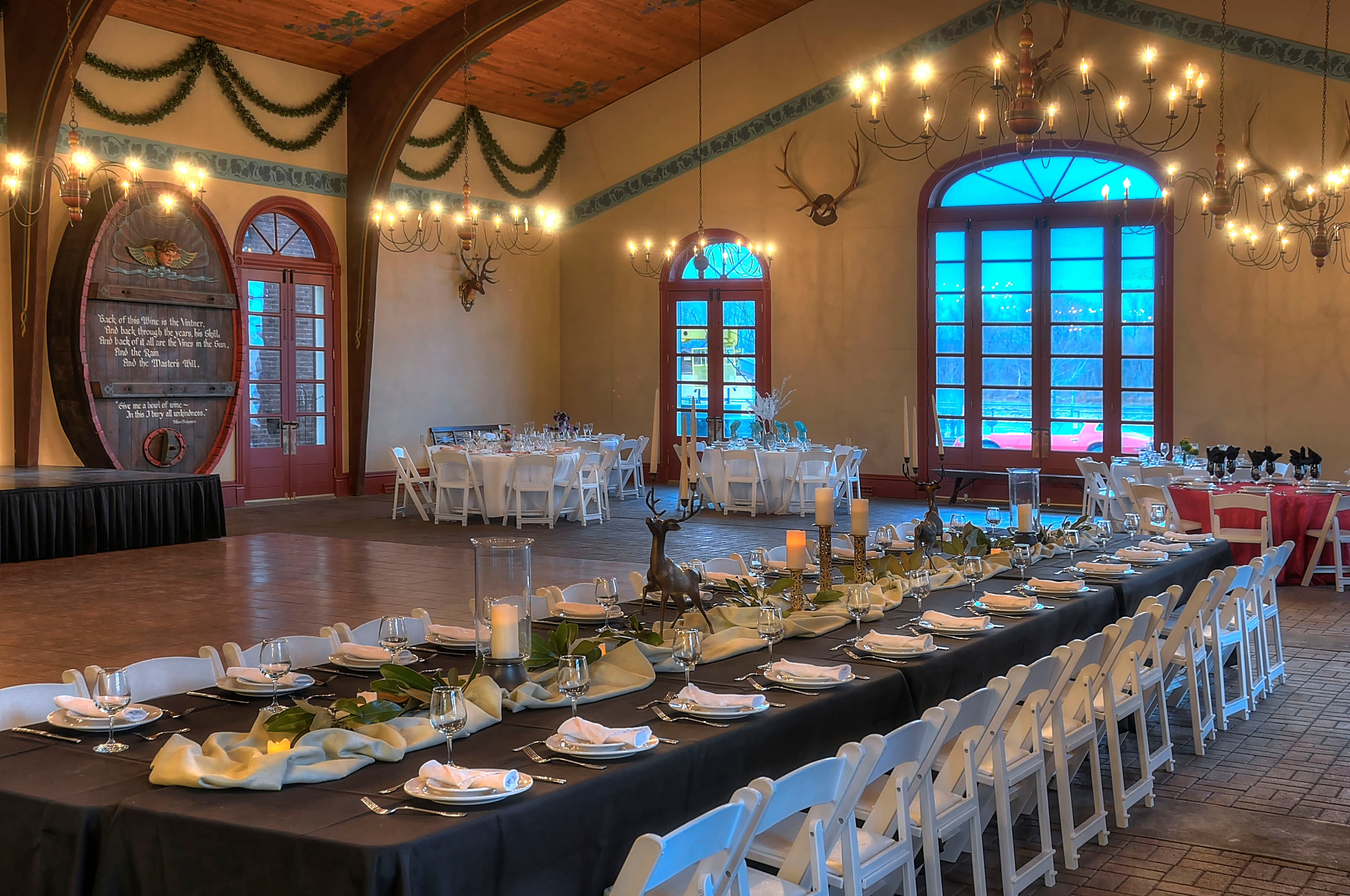 An elegantly set dining area with a long table and white chairs, under chandeliers and next to large windows.
