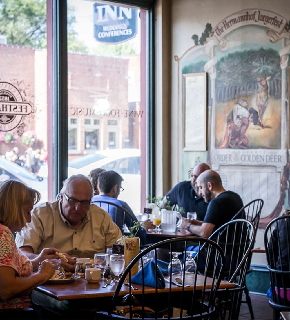 A cozy café scene with patrons enjoying meals at tables near a large window and a mural on the wall.