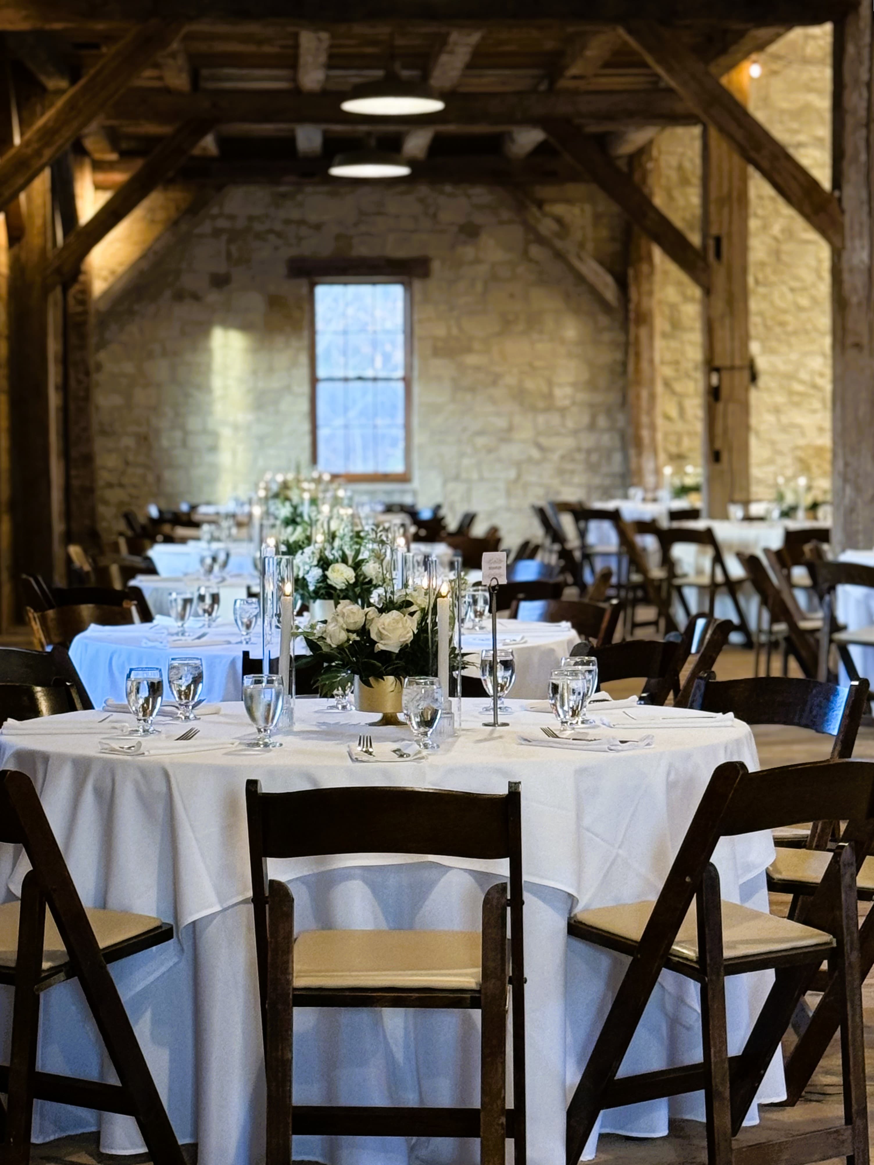 A beautifully set dining area with white tablecloths and flower centerpieces in a rustic venue.