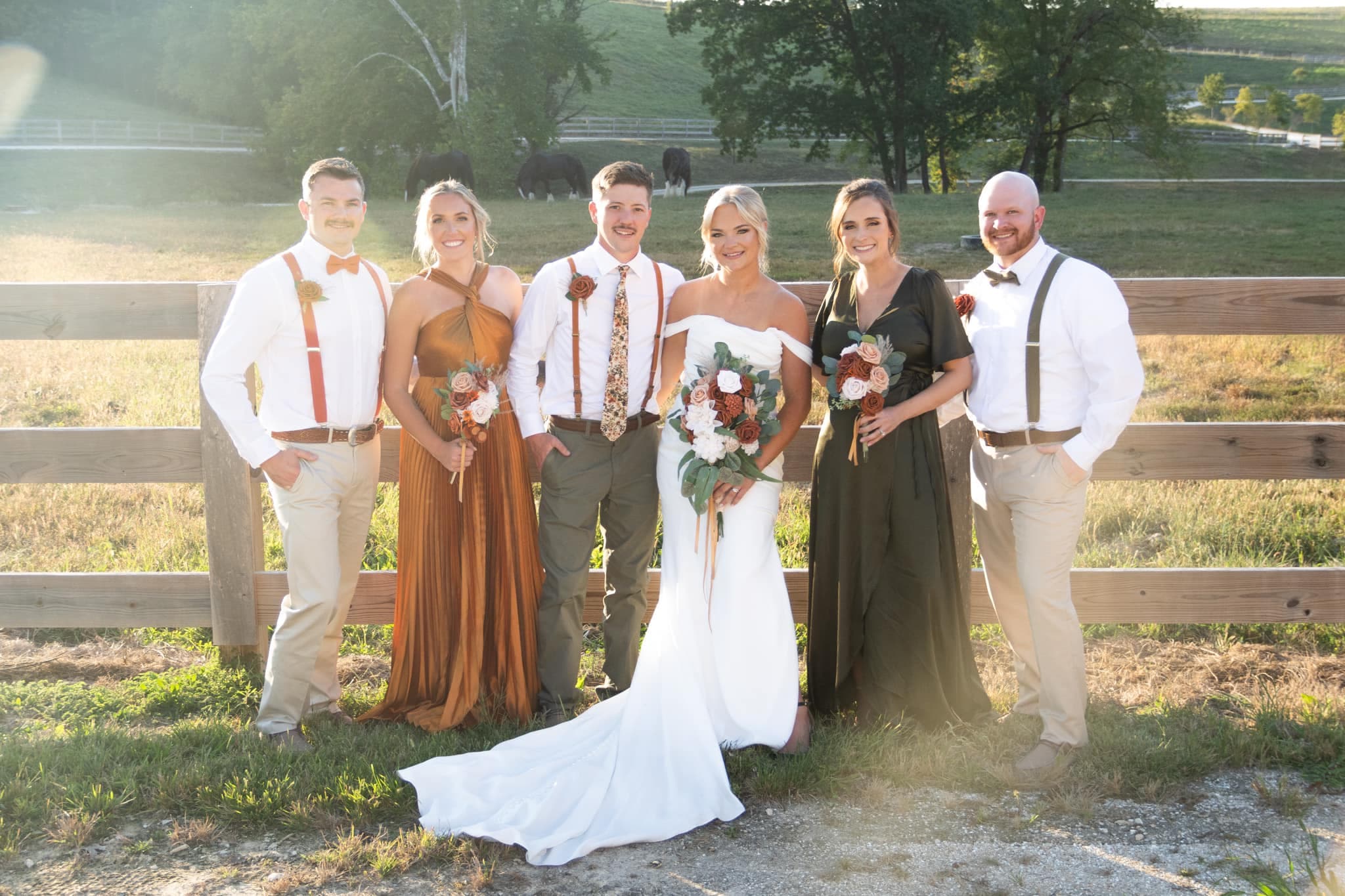 A bride and five wedding attendants stand together outside near a wooden fence, holding bouquets against a scenic backdrop.