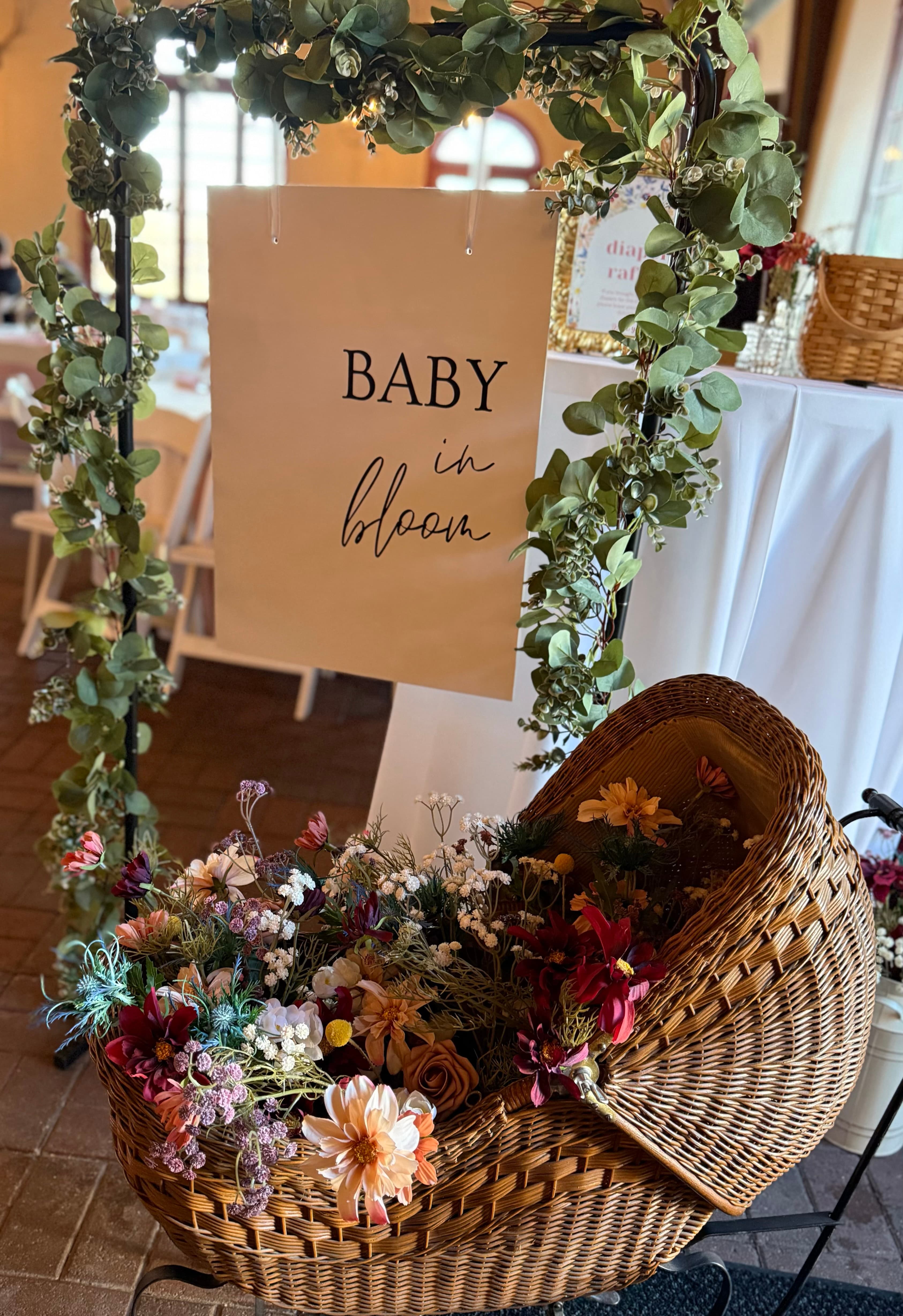 A decorative sign reads "Baby in bloom" surrounded by greenery and a basket filled with colorful flowers.