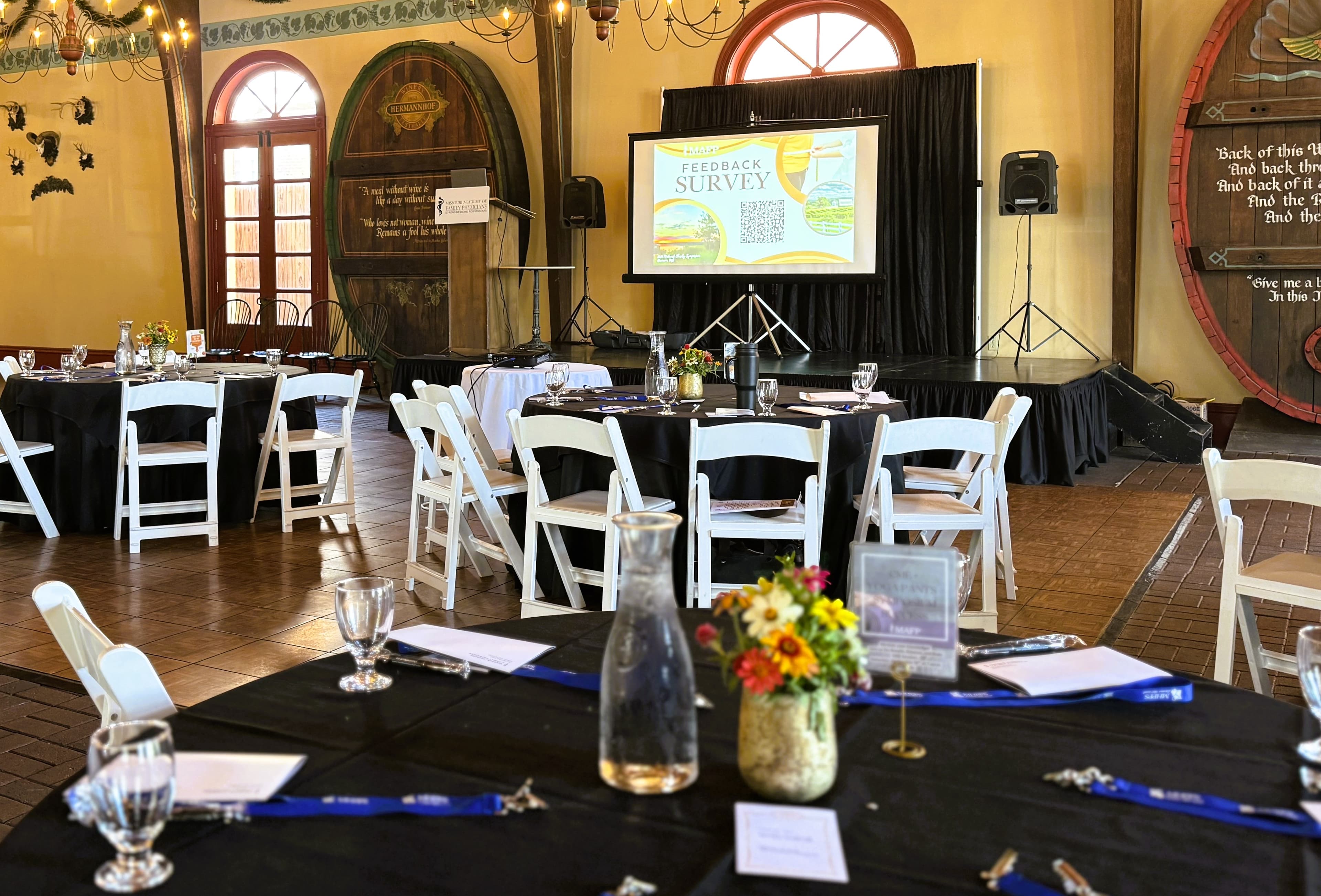 A banquet hall set up for an event, featuring tables with black tablecloths, floral centerpieces, and a presentation screen displaying a feedback survey.