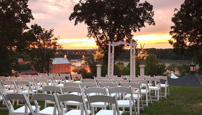 Outdoor wedding ceremony setup with white chairs facing an altar, overlooking a sunset landscape.