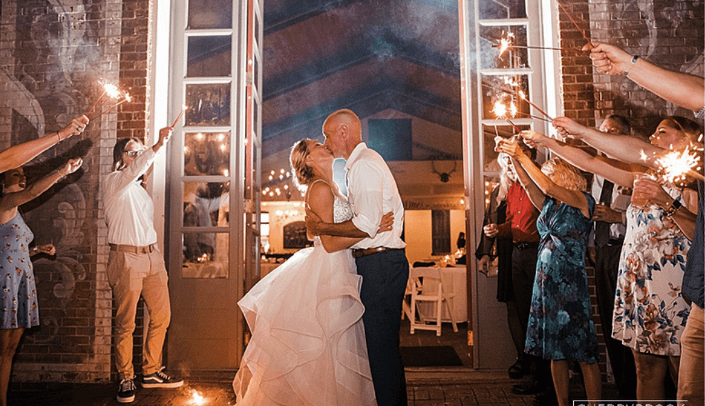 A bride and groom kiss while surrounded by guests holding sparklers in a festive outdoor setting.