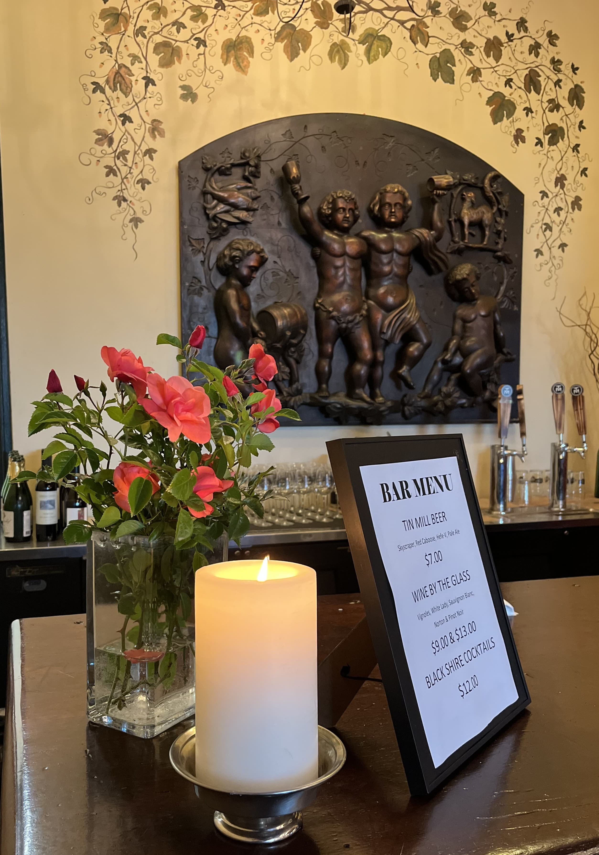 A bar counter featuring a menu, a lit candle, and a vase of pink flowers against a decorative wall with a bronze relief sculpture.