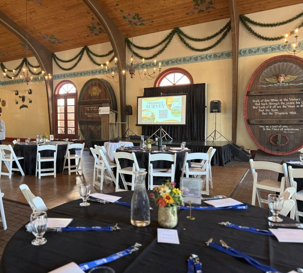 A spacious event venue featuring black tables, white chairs, and a presentation screen, decorated with greenery and warm lighting.