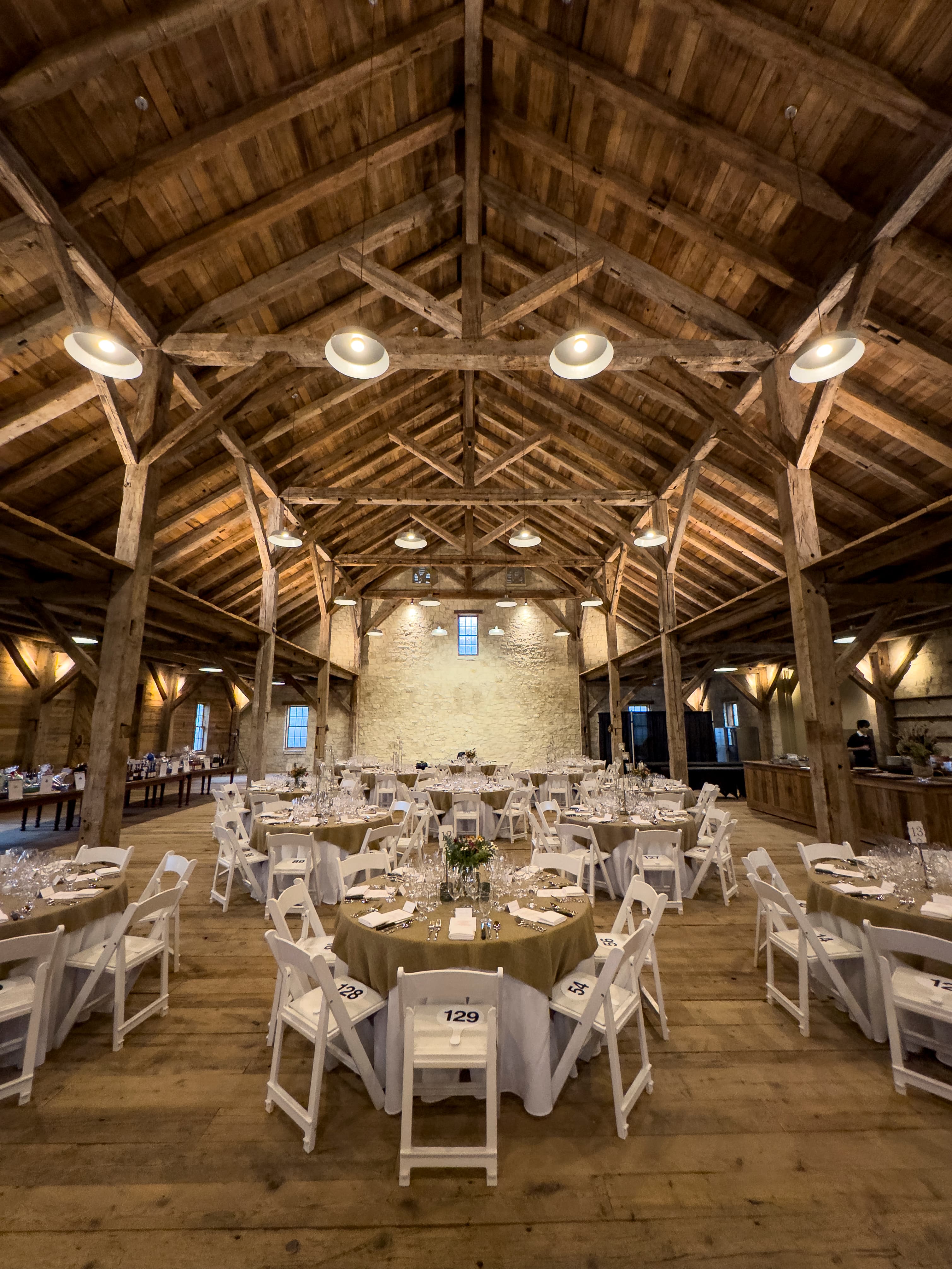 A spacious, rustic event venue featuring round tables set for dining under wooden beams and warm lighting.