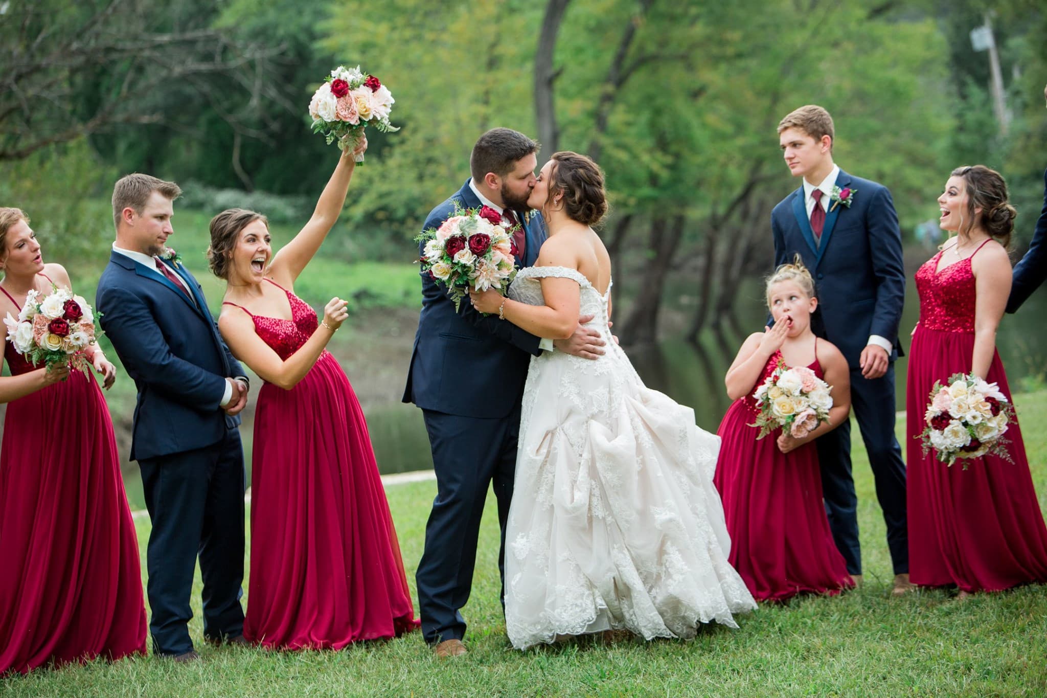 A bride and groom kiss surrounded by their wedding party in coordinating burgundy and navy attire.
