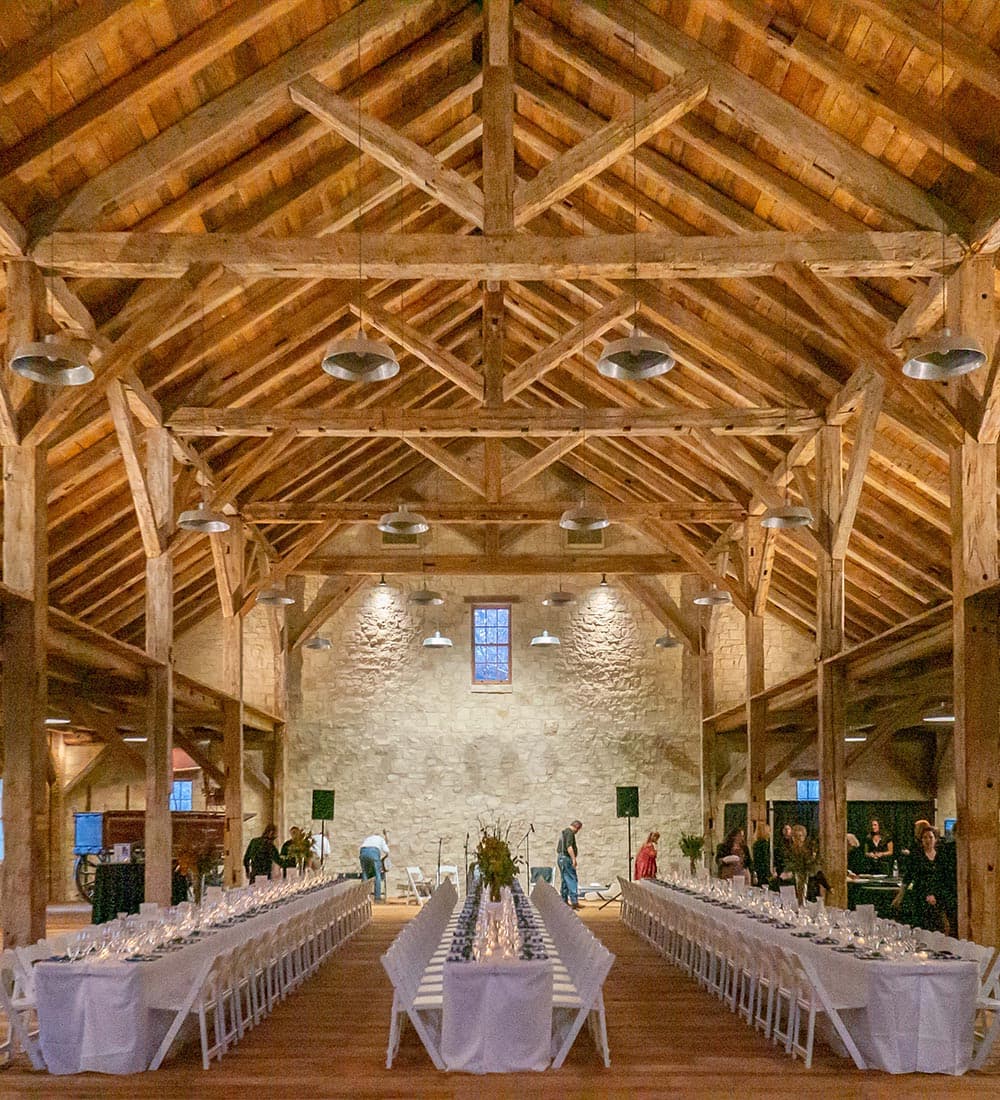 A spacious, elegantly decorated barn interior featuring long tables set for an event under a wooden beam ceiling.